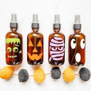 May include: Four brown glass bottles with different Halloween-themed designs. The bottles have black lids and are filled with a clear liquid. The designs include a pumpkin, a monster, a mummy, and a ghost.