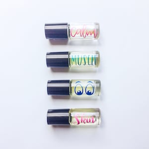 May include: Four small glass bottles with black roll-on tops. The bottles are filled with a clear liquid and labeled with different words: "Calm", "Muscle", "Eyes", and "Skin".