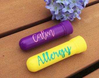 May include: Two small, labeled tubes, one purple with the word "calm" and one yellow with the word "allergy", on a wooden surface. A cluster of light blue flowers is in the background.