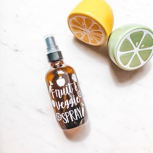 May include: A brown glass spray bottle with a white label that says "Fruit & Veggie Spray". The bottle is sitting on a white surface with two decorative lemons.
