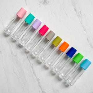 Puede incluir: Diez botellas de vidrio con rodillo y tapas de plástico de colores. Las tapas son rosa, verde azulado, lavanda, rosa intenso, gris, verde oliva, naranja, azul, verde lima y azul claro.