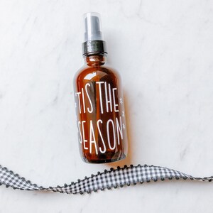 May include: A brown glass bottle with a black spray top and a white label that says "Tis the Season" with snowflakes.