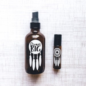May include: Two amber glass bottles with black lids and white labels. The larger bottle has a spray nozzle and the label says "Dream Big" with a dreamcatcher design. The smaller bottle has a rollerball applicator and a dreamcatcher design on the label.