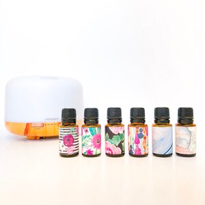 May include: A white and wood essential oil diffuser with six small amber glass bottles with colorful floral and geometric patterned labels.  The labels include the text "LIGHT", "ON", "1H", "2H", "CONTINOUS", and "TIMER".