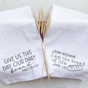 Puede incluir: Dos toallas de cocina blancas con texto negro. La primera toalla dice "Danos hoy nuestro pan de cada día Mateo 6:11". La segunda toalla dice "Jesús declaró. Yo soy el pan de vida. Juan 6:35".