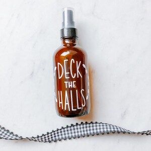May include: A brown glass bottle with a clear spray nozzle. The bottle has a white label with the text "Deck the Halls" in a handwritten font.