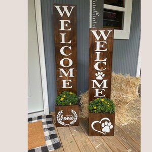 May include: Two wooden welcome signs with white lettering and a dark brown stain. One sign has a paw print and heart design, the other has a wreath design. Both have a planter box with greenery.