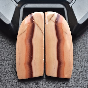 Puede incluir: Dos cabujones pulidos en forma de semicírculo de piedra veteada con capas verticales en tonos beige, marrón claro y marrón chocolate oscuro. Las piedras presentan un acabado liso y brillante con vetas naturales visibles y definidas.