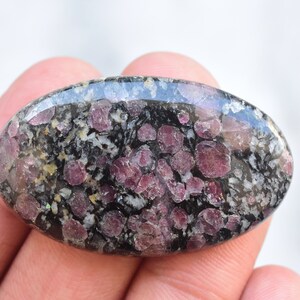 K&ouml;nnte beinhalten: Oval geformter Edelstein mit polierter Oberfl&auml;che. Der Stein weist ein gesprenkeltes Muster aus Dunkelgrau, Wei&szlig; und tiefroten Farbt&ouml;nen auf. Die roten Flecken sind &uuml;ber den gesamten Stein verteilt und erzeugen eine einzigartige visuelle Textur. Der Edelstein wird in einer Hand gehalten.