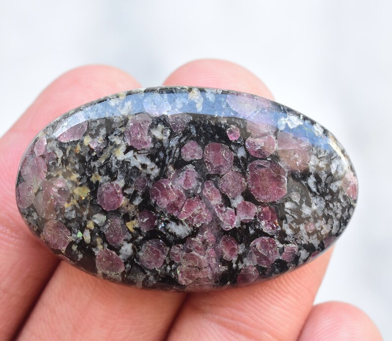 K&ouml;nnte beinhalten: Oval geformter Edelstein mit polierter Oberfl&auml;che. Der Stein weist ein gesprenkeltes Muster aus Dunkelgrau, Wei&szlig; und tiefroten Farbt&ouml;nen auf. Die roten Flecken sind &uuml;ber den gesamten Stein verteilt und erzeugen eine einzigartige visuelle Textur. Der Edelstein wird in einer Hand gehalten.