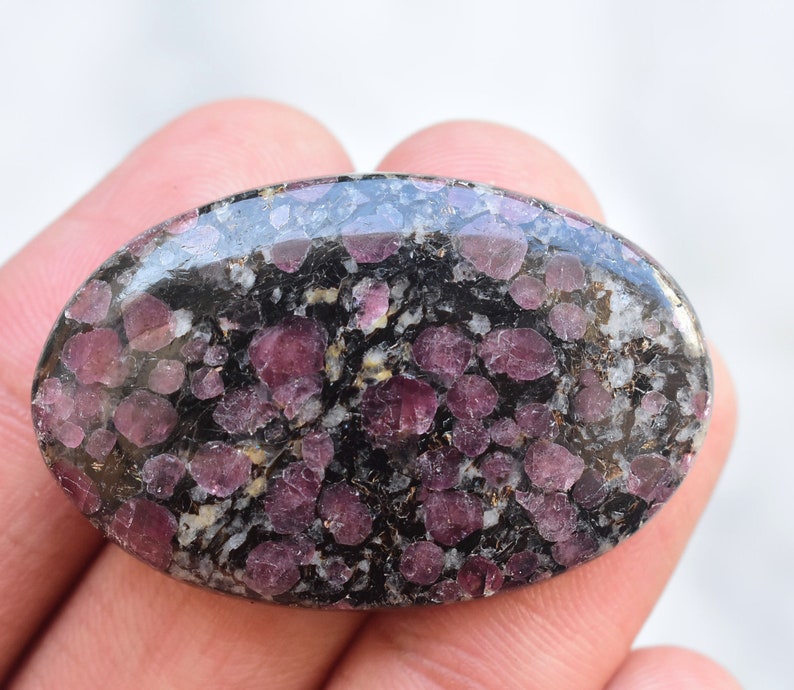 Op de afbeelding: Een ovaalvormige gepolijste edelsteen cabochon met een donkerzwarte matrix ingebed met talrijke afgeronde, diep magenta en paars getinte minerale insluitingen over het hele oppervlak, wat zorgt voor een contrastrijk gespikkeld uiterlijk.