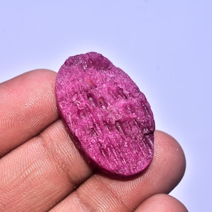 Könnte beinhalten: Ein rauer, oval geformter, tiefrosa Rubin-Edelstein. Die Oberfläche hat ein strukturiertes, unebenes Aussehen mit sichtbaren Rippen und Rissen. Der Edelstein wird in einer Hand gehalten.