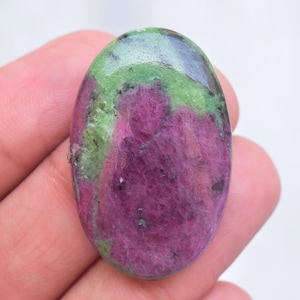 Puede incluir: Una piedra preciosa pulida de forma ovalada que presenta un centro de color magenta vibrante rodeado de inclusiones minerales de color verde lima y verde bosque oscuro. La superficie de la piedra es lisa y reflectante, mostrando variaciones de color naturales.