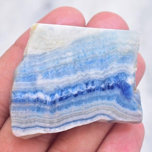 May include: A rectangular piece of blue calcite stone with layered bands of white, light blue, and deep blue hues. The stone has a rough, natural edge and a polished surface, showcasing the mineral's unique patterns.