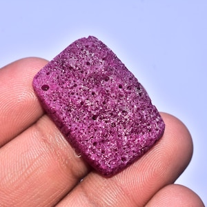 Könnte beinhalten: Ein rechteckiger, poröser, magentafarbener Edelstein mit einem kleinen Loch oben. Der Stein hat eine raue, strukturierte Oberfläche und wird in einer Hand vor einem hellblauen Hintergrund gehalten.