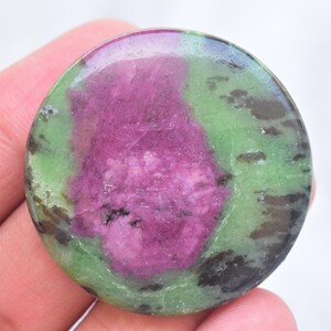 Peut inclure: Une pierre précieuse de rubis zoïsite polie et circulaire, présentant un centre magenta-rose vif entouré d'une matrice vert clair tachetée avec des inclusions noires irrégulières sur toute la surface lisse et réfléchissante.