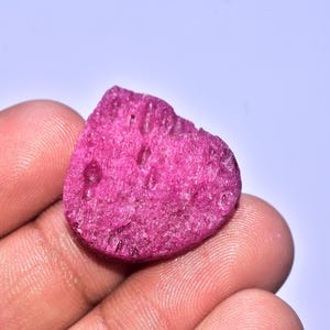 Könnte beinhalten: Ein grob geschnittener, herzförmiger, fuchsienfarbener Edelstein. Der Stein hat eine strukturierte Oberfläche mit kleinen Löchern und wird in der Hand einer Person vor einem hellvioletten Hintergrund gehalten.
