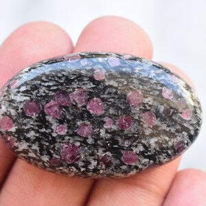 K&ouml;nnte beinhalten: Ein oval geschliffener Edelstein mit einer schwarz-wei&szlig; gesprenkelten Matrixbasis, in die zahlreiche kleine, abgerundete, leuchtend magentafarbene Kristalle eingebettet sind, die &uuml;ber die glatte, reflektierende Oberfl&auml;che verteilt sind.