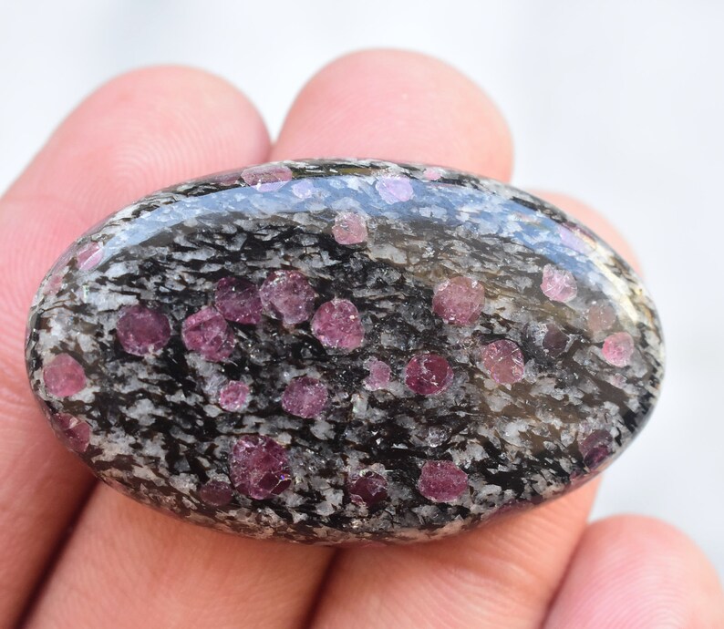 K&ouml;nnte beinhalten: Ein oval geschliffener Edelstein mit einer schwarz-wei&szlig; gesprenkelten Matrixbasis, in die zahlreiche kleine, abgerundete, leuchtend magentafarbene Kristalle eingebettet sind, die &uuml;ber die glatte, reflektierende Oberfl&auml;che verteilt sind.