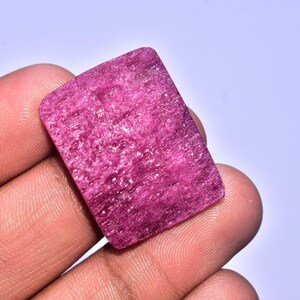 Pode incluir: Uma pedra preciosa retangular, rosa escuro, com textura áspera. A pedra é segurada na mão, mostrando sua superfície natural e não polida. A cor é um rosa vibrante e saturado.
