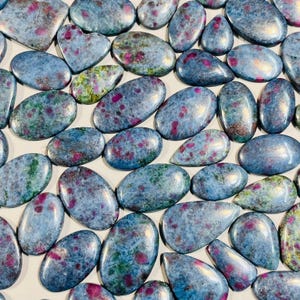 May include: A collection of polished cabochons featuring a blue kyanite base with embedded red ruby inclusions. These smooth, rounded stones display a mix of oval, teardrop, and heart shapes, showcasing natural color variations of blue, red, and green.