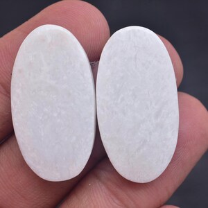 May include: Two oval-shaped, smooth, white gemstones with a subtle, cloudy, and marbled texture throughout their surfaces. These cabochons feature a polished finish and a consistent, opaque appearance, showcasing a soft, milky white colour palette.