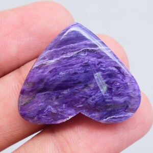 Puede incluir: Una piedra preciosa pulida en forma de corazón, con un patrón arremolinado de púrpura intenso y blanco. La piedra tiene una superficie lisa y brillante y una textura natural y orgánica. La piedra se sostiene en una mano.