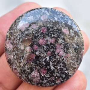 Könnte beinhalten: Ein runder, polierter Stein mit einem gesprenkelten schwarz-grauen Hintergrund. Der Stein ist mit zahlreichen kleinen, runden, tiefrosa Edelsteinen übersät. Die Oberfläche ist glatt und reflektierend, mit leichtem Glanz.