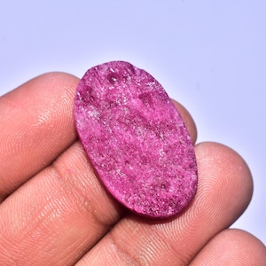 May include: A rough, oval-shaped ruby gemstone with a deep pink hue. The gemstone is held in a hand, showcasing its textured surface and natural imperfections. The ruby is a vibrant, eye-catching piece.