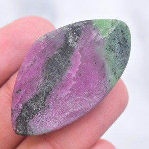 May include: A polished marquise-shaped ruby zoisite gemstone featuring a vibrant mix of magenta, deep forest green, and charcoal black mineral inclusions. The stone has a smooth, flat surface with a natural, irregular color distribution throughout.