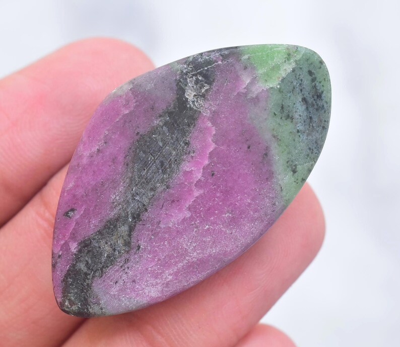 May include: A polished marquise-shaped ruby zoisite gemstone featuring a vibrant mix of magenta, deep forest green, and charcoal black mineral inclusions. The stone has a smooth, flat surface with a natural, irregular color distribution throughout.