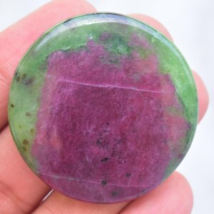 May include: A circular polished gemstone featuring a vibrant magenta-pink center surrounded by a smooth, translucent light green border. The stone has a flat, disc-like shape with visible natural mineral inclusions and a glossy, reflective surface finish.