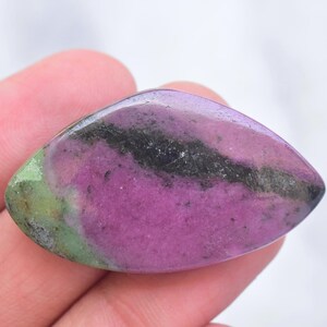 May include: A polished marquise-shaped gemstone featuring a vibrant magenta-pink center, a distinct dark black mineral inclusion running horizontally across the surface, and a soft lime-green accent on the left side with a smooth, glossy finish.