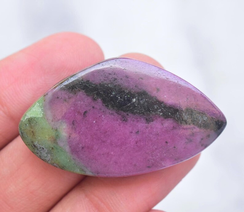 May include: A polished marquise-shaped gemstone featuring a vibrant magenta-pink center, a distinct dark black mineral inclusion running horizontally across the surface, and a soft lime-green accent on the left side with a smooth, glossy finish.