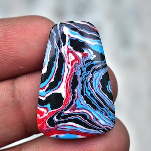 May include: A rectangular gemstone with a distinctive swirling pattern. The design incorporates red, white, black, and blue streaks, producing a marbled effect. The stone is held in a hand, highlighting its polished surface and vibrant colour palette.