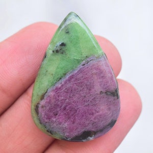 May include: A polished, teardrop-shaped ruby zoisite gemstone featuring a distinct diagonal color split. The stone displays a vibrant, opaque lime green section contrasted against a deep magenta-pink area, with small black mineral inclusions.