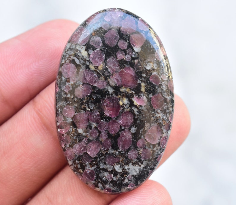 Op de afbeelding: Een ovaalvormige gepolijste edelsteen cabochon met een donkerzwarte en grijze matrix, ingebed met talrijke afgeronde, doorschijnende dieproze tot magentakleurige minerale insluitingen die over het gehele oppervlak van de steen zijn verdeeld.