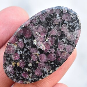 K&ouml;nnte beinhalten: Ein ovaler Edelstein mit einem gesprenkelten Muster. Der Stein hat eine dunkelgraue Basis mit Clustern aus tiefroten und rosa Kristallen. Die polierte Oberfl&auml;che reflektiert das Licht und hebt die einzigartige Textur und Farbvariationen des Natursteins hervor.