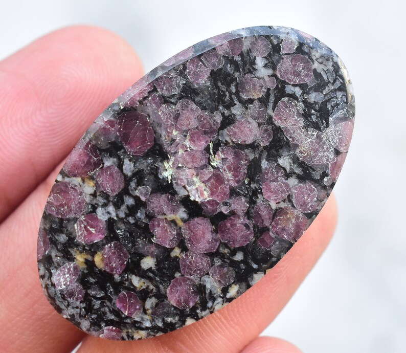 K&ouml;nnte beinhalten: Ein ovaler Edelstein mit einem gesprenkelten Muster. Der Stein hat eine dunkelgraue Basis mit Clustern aus tiefroten und rosa Kristallen. Die polierte Oberfl&auml;che reflektiert das Licht und hebt die einzigartige Textur und Farbvariationen des Natursteins hervor.