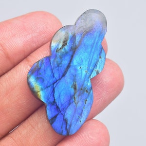 Pode incluir: Uma pedra preciosa de labradorite polida com uma forma irregular distinta semelhante a uma nuvem. A pedra exibe uma vibrante labradorescência azul iridescente na sua superfície, acentuada por finas inclusões e veios naturais escuros.