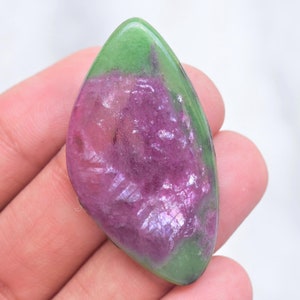 May include: A polished marquise-shaped cabochon gemstone featuring a vibrant magenta-pink center surrounded by a smooth, opaque green border. The surface of the stone displays a glossy, reflective finish with natural crystalline textures visible.