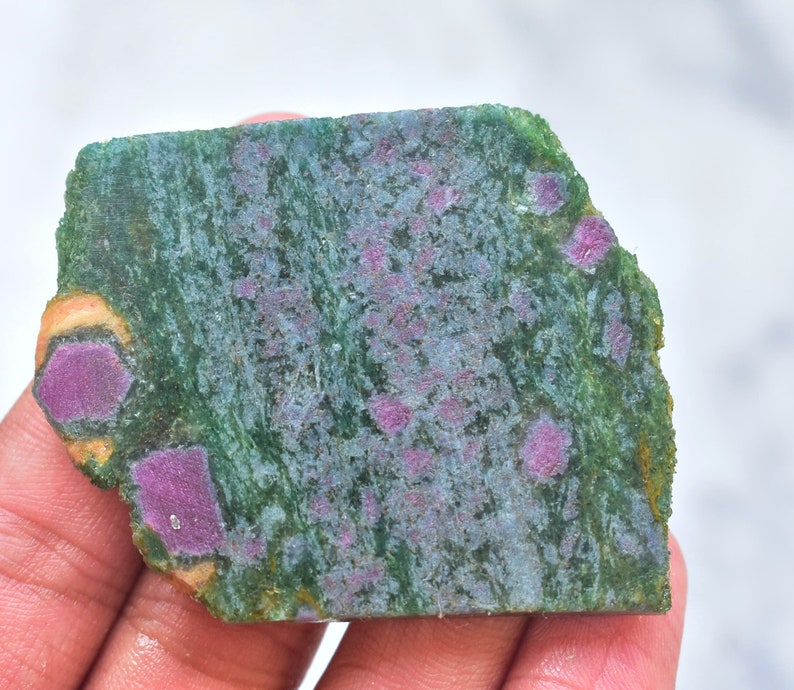 K&ouml;nnte beinhalten: Eine flache, unregelm&auml;&szlig;ig geformte Platte aus gr&uuml;nem Zoisit-Mineral mit eingebetteten, leuchtend magentafarbenen Rubinkristallen. Die Oberfl&auml;che zeigt ein strukturiertes, gesprenkeltes Aussehen mit tiefgr&uuml;nen Streifen und deutlichen, opaken rot-rosa Einschl&uuml;ssen.