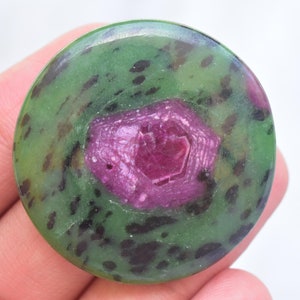May include: A circular polished gemstone cabochon featuring a vibrant magenta-pink hexagonal crystal inclusion centered within a mottled green and black zoisite matrix. The surface is smooth and rounded, displaying natural color variations.