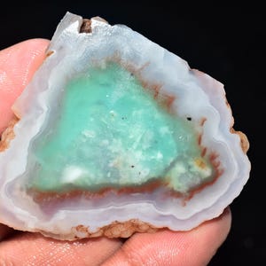 May include: A polished gemstone slice with a vibrant turquoise center, surrounded by bands of white, brown, and red. The stone has a rough, natural edge and is held in a hand against a black background. This unique piece showcases natural mineral formations.
