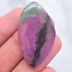May include: A polished, marquise-shaped ruby zoisite gemstone featuring a vibrant magenta-pink center, deep green edges, and a prominent dark black mineral inclusion running vertically through the right side of the smooth, reflective surface.