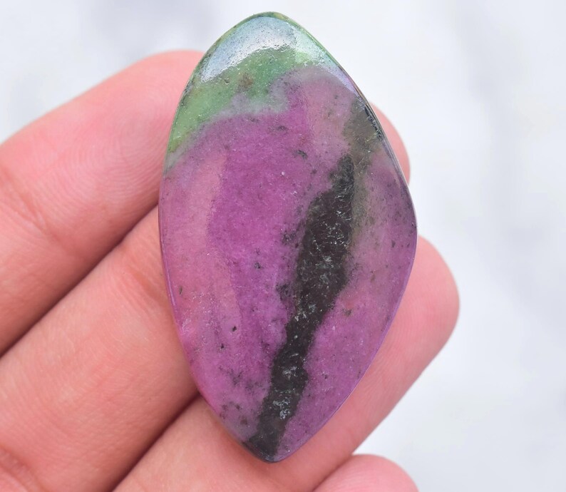 May include: A polished, marquise-shaped ruby zoisite gemstone featuring a vibrant magenta-pink center, deep green edges, and a prominent dark black mineral inclusion running vertically through the right side of the smooth, reflective surface.