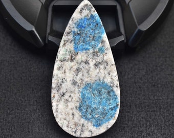 May include: A teardrop-shaped cabochon gemstone featuring a speckled white, gray, and black granite-like matrix with two prominent, vibrant blue circular inclusions. The polished stone has a smooth surface and a pointed top with a rounded base.