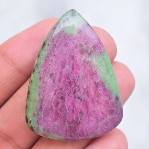 Op de afbeelding: Een gepolijste, druppelvormige robijn-zoisiet edelsteen cabochon met een levendig magenta-roze centrum, omringd door een gladde, lichtgroene rand met kleine donkere insluitingen over het gehele oppervlak van de steen.