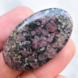 K&ouml;nnte beinhalten: Ein ovaler Edelstein mit polierter Oberfl&auml;che. Der Stein hat einen dunklen Hintergrund, der mit Wei&szlig; und Grau gesprenkelt ist, und Ansammlungen von tiefroten, runden Einschl&uuml;ssen. Der Edelstein wird in einer Hand gehalten.