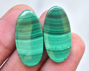 Puede incluir: Dos piedras preciosas ovaladas de malaquita pulida. Las piedras muestran un color verde vibrante con patrones de bandas verdes y marrones más oscuros. Las piedras preciosas son lisas y tienen un acabado brillante.